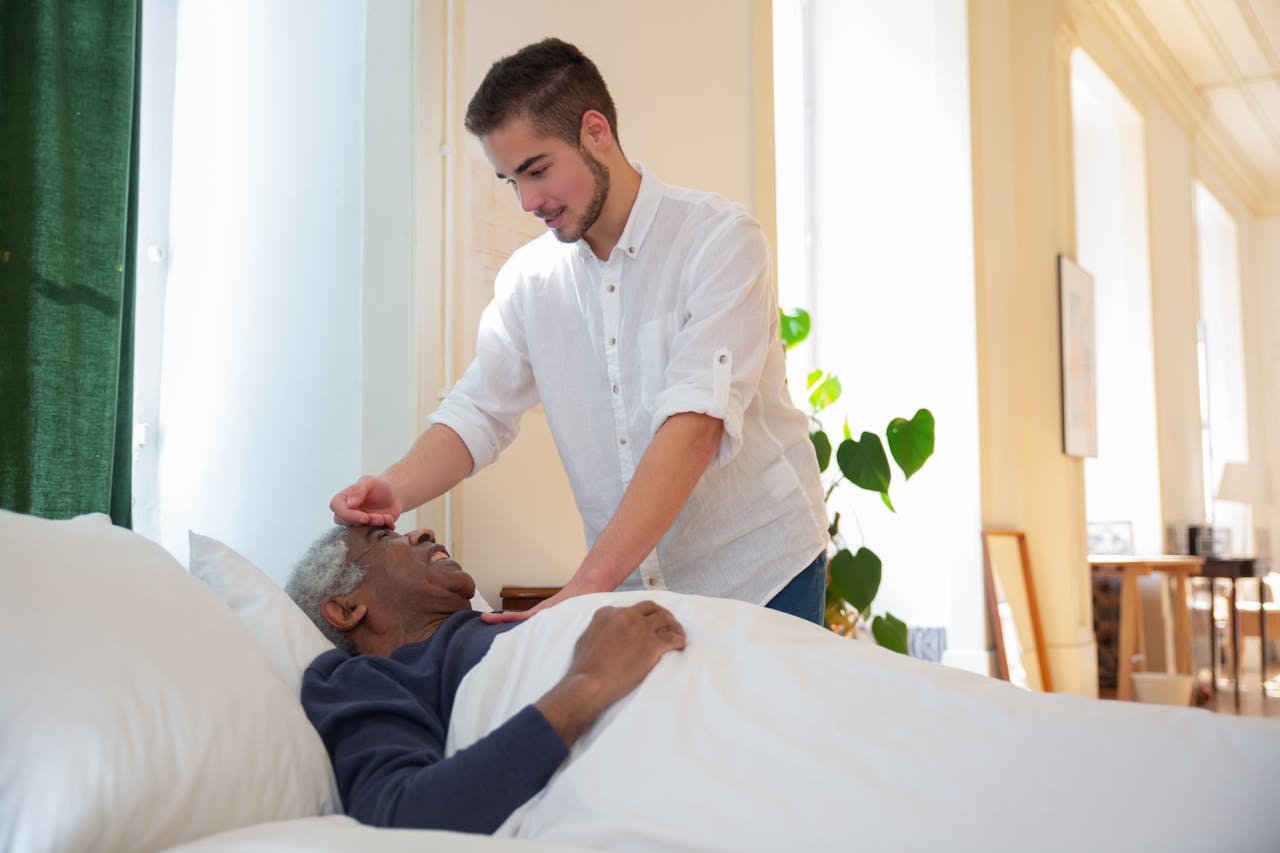 A young man supports an elderly man in bed, offering care in a bright room.